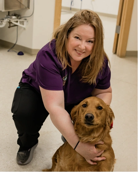 A staff member in a purple shirt kneeling beside a golden retriever, smiling at the camera.