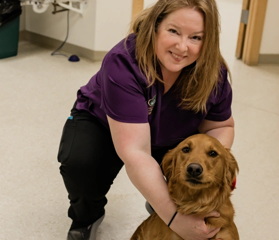 Veterinary staff member in purple scrubs kneeling and hugging a golden retriever in the clinic hallway